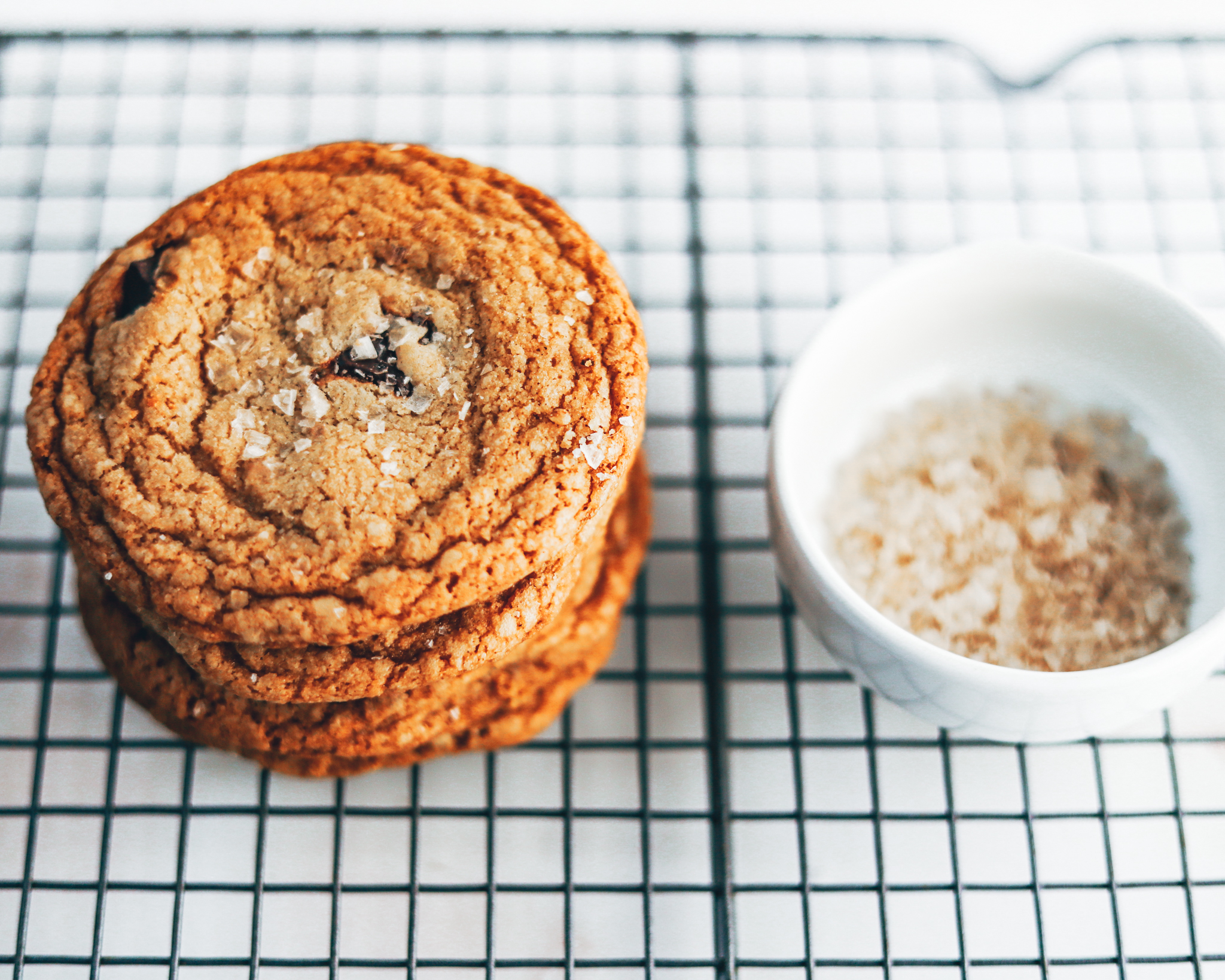 Stacked chocolate chip cookies sit on a cooling rack next to a bowl of flakey sea salt for garnishing