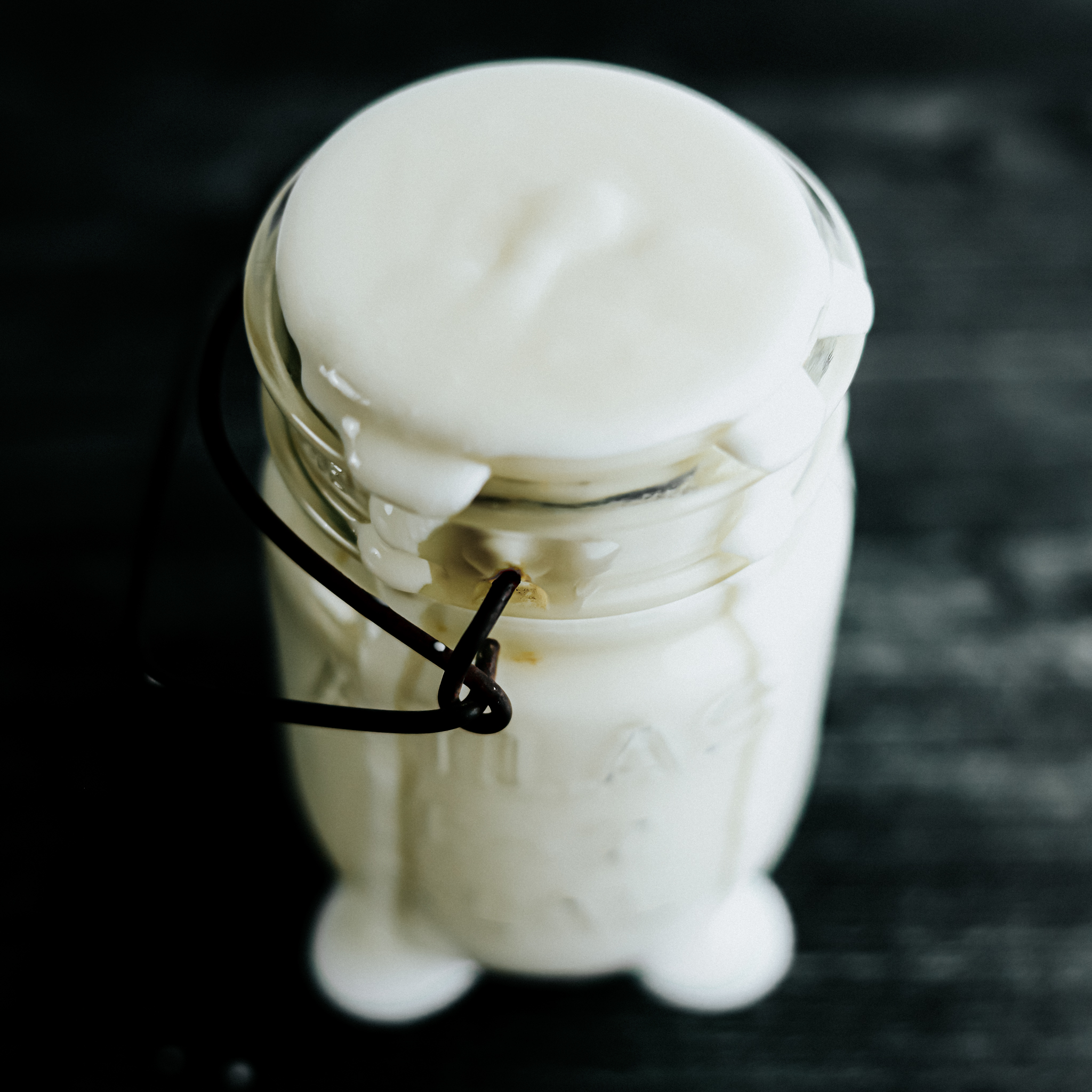 Vintage glass jar with rust iron hinge holds a fresh batch of cultivated kefir, slightly overflowing from the top and onto the gray wooden countertops