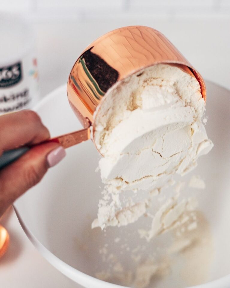 hand pours a rose gold measuring cup of flour into a white bowl