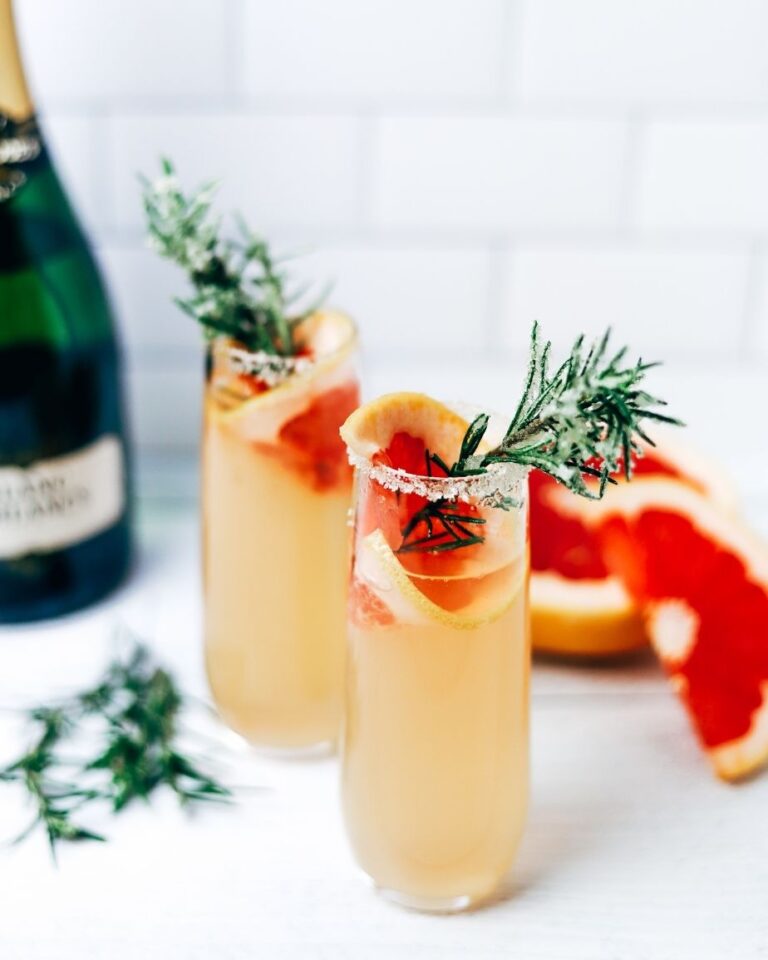 two champagne glasses filled with light orange cocktail and garnished with a bright orange grapefruit slice and a sprig of rosemary
