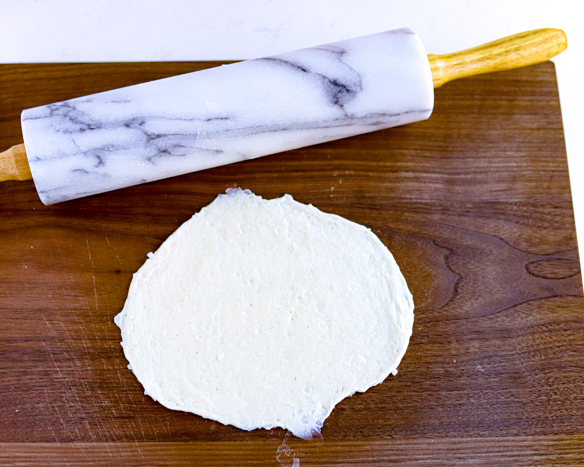 biscuit dough rolled out into a circle on to of wooden cutting board with grey marbled rolling pin the background