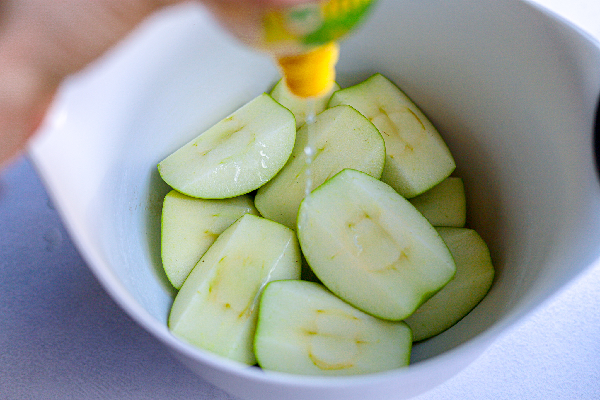 Bowl of green Granny Smith apples cut into quarters. A bottle of lemon juice hovers above, squeezing juice onto the apples.
