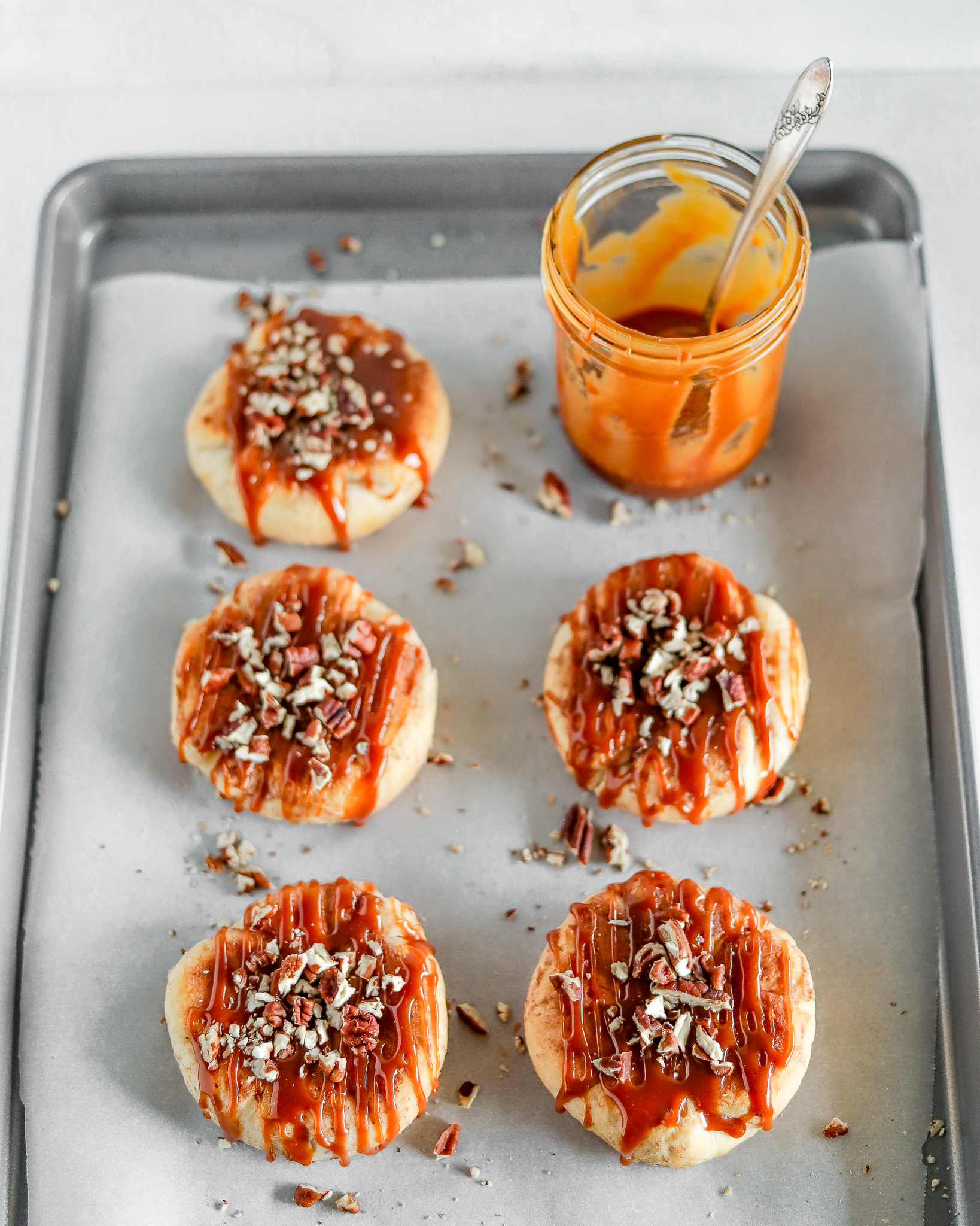 overhead view of sheet pan with two rows of caramel apple biscuits topped with chopped pecans and a jar of caramel in the background