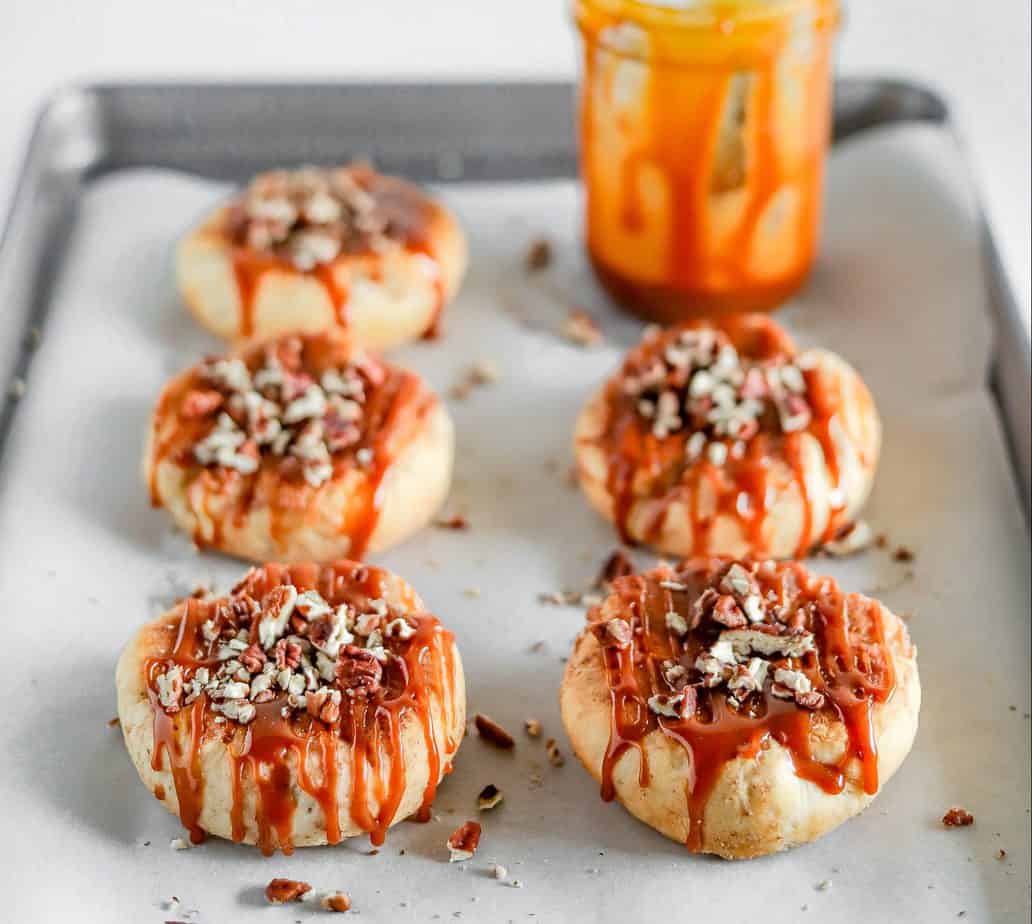 5 caramel apple biscuits topped with chopped walnuts arranged in two rows on a baking sheet with a jar of half empty caramel in the background. spoon in the caramel jar with caramel dripping down the side of the jar.
