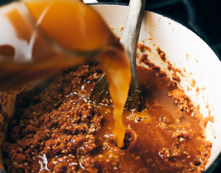 chicken broth pours from a pyrex measuring cup into a pot of simmering bolognese