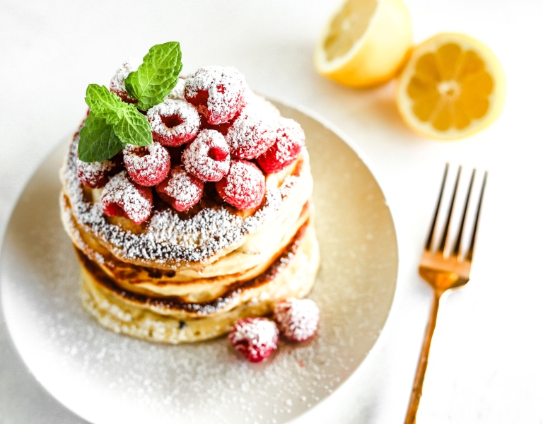 stack of lemon ricotta pancakes with raspberries and powdered sugar; rose gold fork on table and lemons in the background